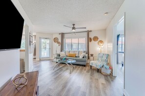 A spacious living room with a grey sofa, a coffee table, and a patterned armchair. A ceiling fan and a window with blinds are visible. - Tierra Verde Bayview Condo | 2BR 2BA (St. Petesburg)