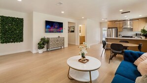 A modern living room with a round table, a sofa, a TV mounted on the wall, and a kitchen in the background. - Sun Chaser | Pool | Spacious Home | Parking (Monterey Park)