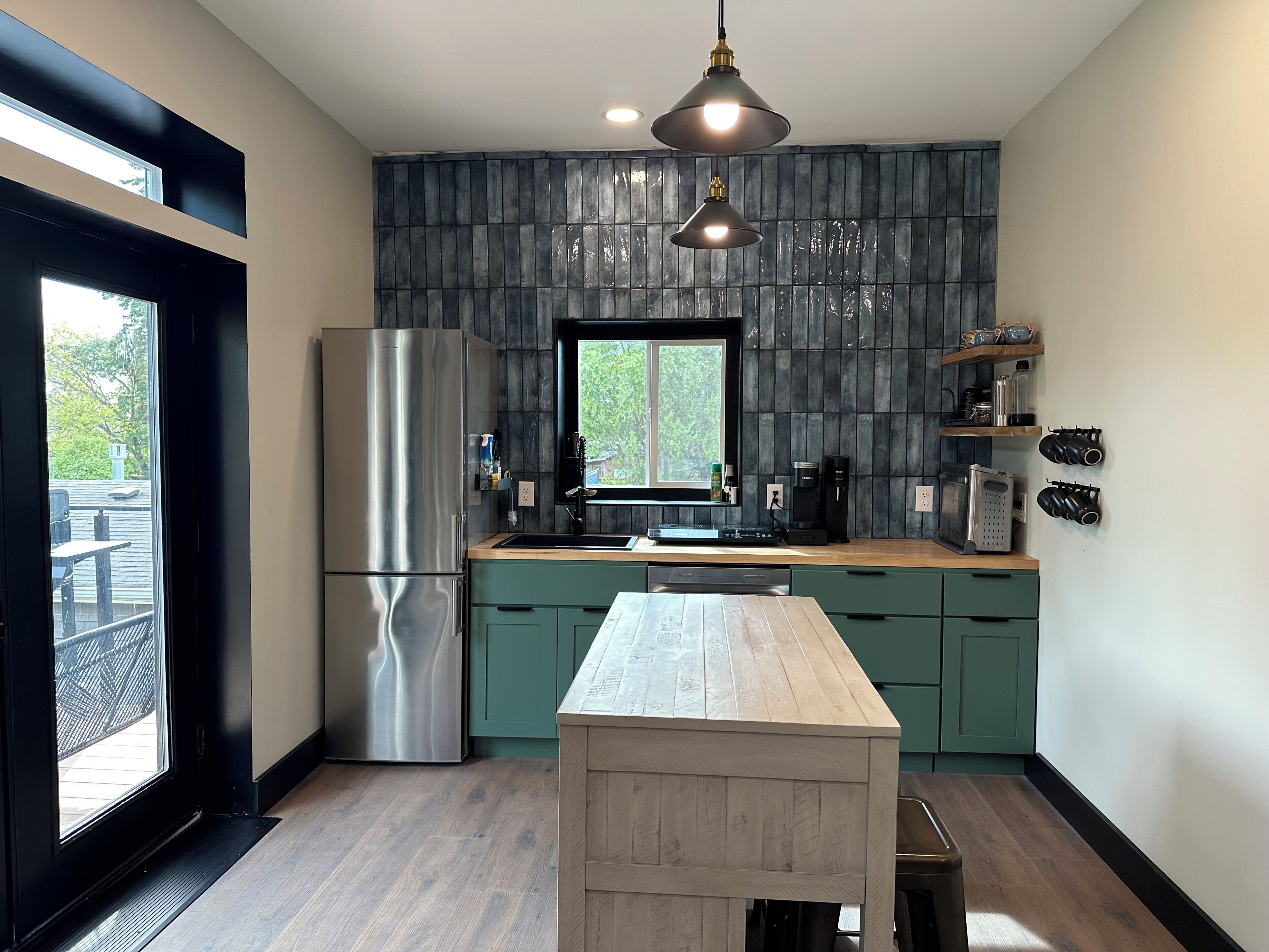 Une cuisine moderne avec un réfrigérateur en acier inoxydable, une table en bois et une fenêtre donnant sur un espace verdoyant.