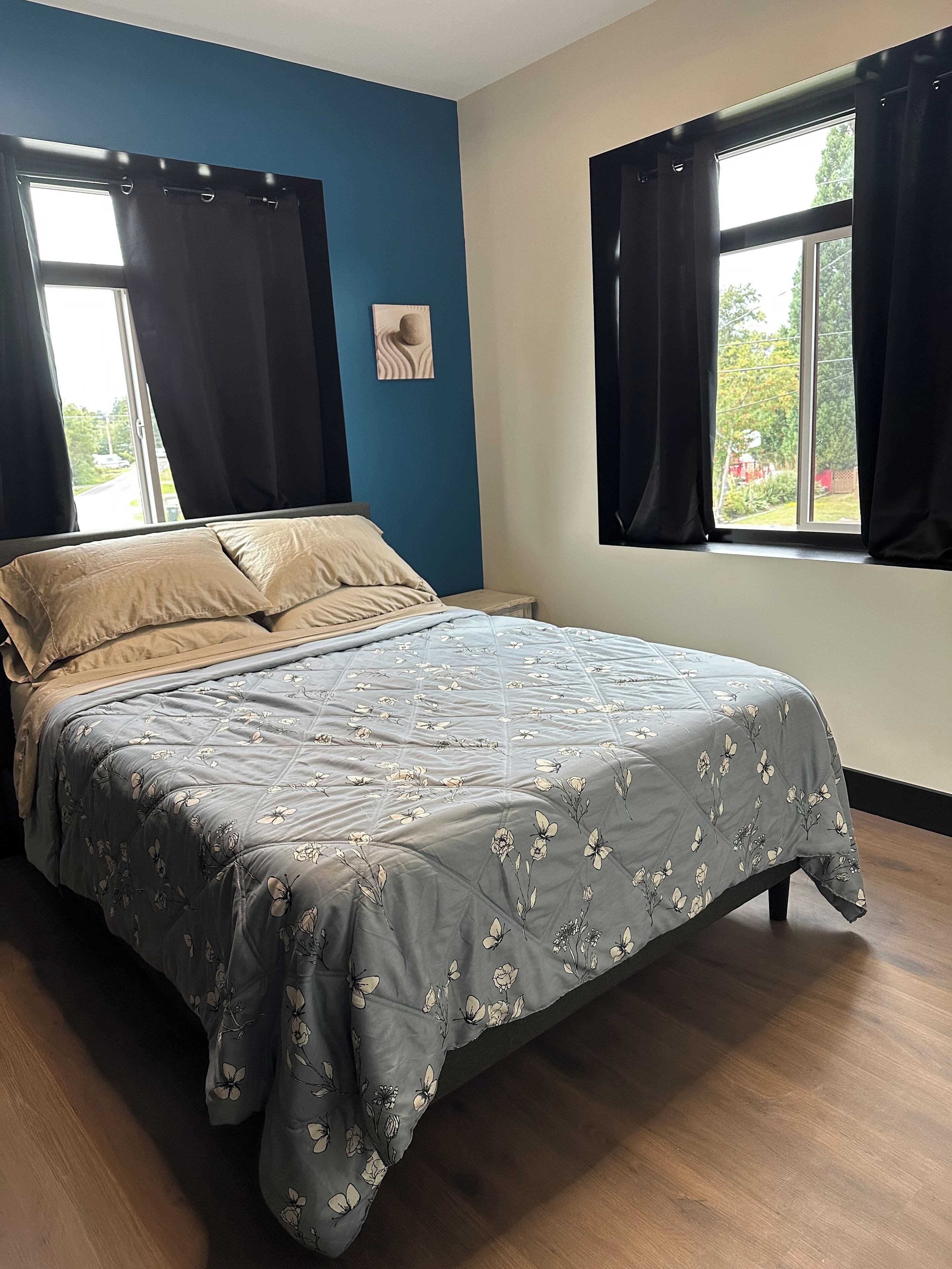 A bedroom with a bed, two windows with curtains, a wall art piece, and a view of greenery outside.