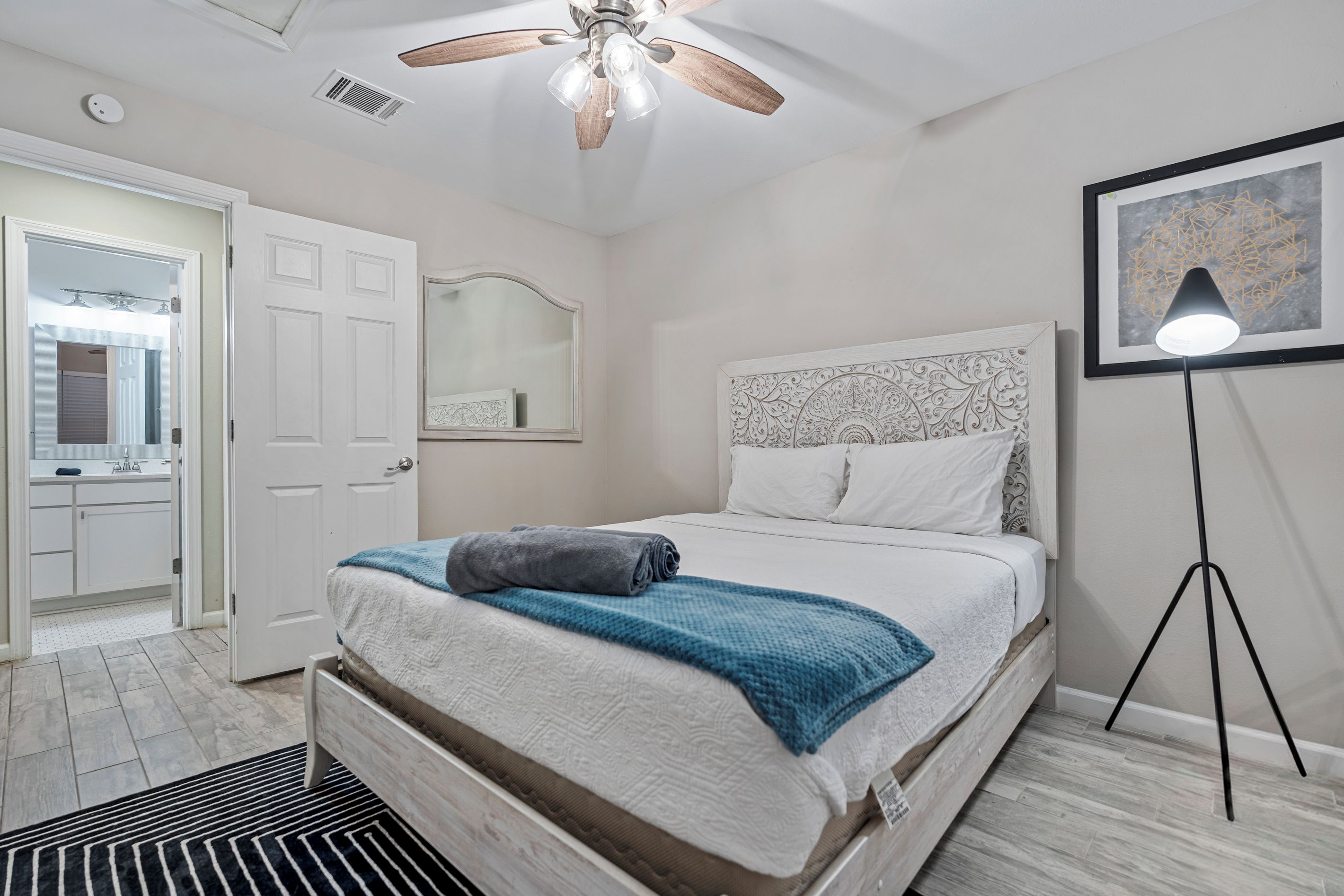 A bedroom with a bed, a ceiling fan, a standing lamp, a mirror, and a door leading to a bathroom.
