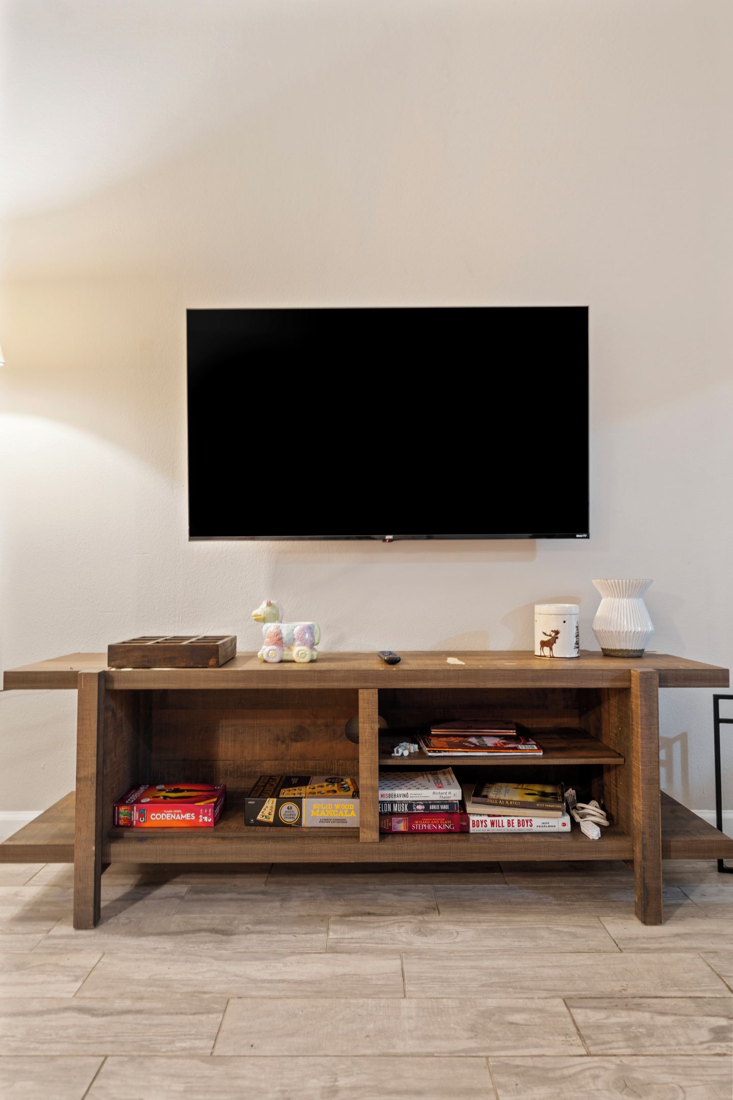 A flat-screen TV mounted on a wall above a wooden entertainment center with shelves containing books and board games.