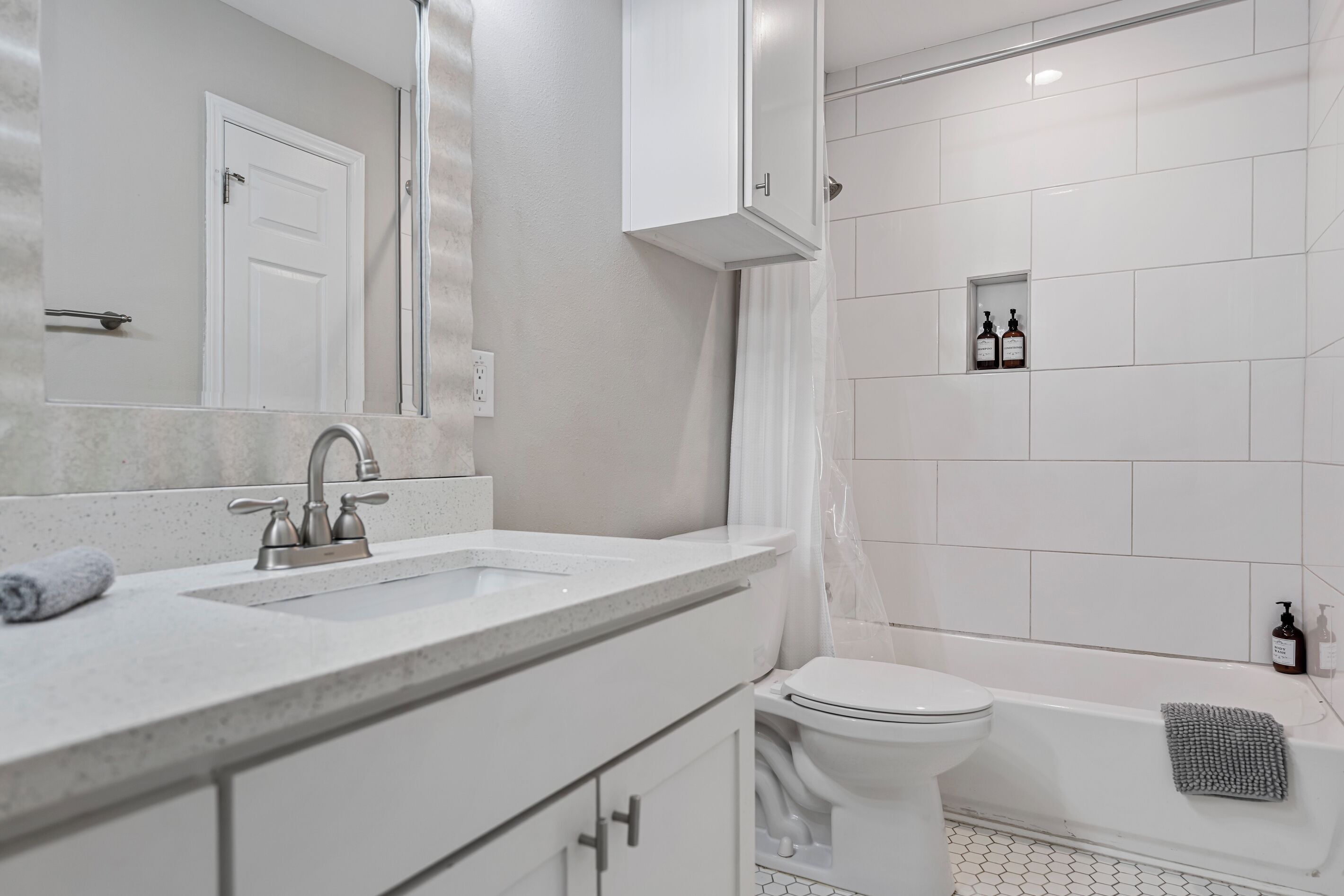 A modern bathroom with a double sink, toilet, and bathtub. There are toiletries in a shelf and a towel on the floor.