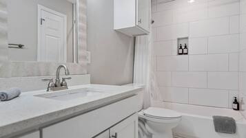 A modern bathroom with a double sink, toilet, and bathtub. There are toiletries in a shelf and a towel on the floor.