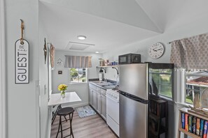 Eine moderne Küche mit weißen Schränken, einem Edelstahlkühlschrank, einer Mikrowelle und einem Herd. An der Wand hängt eine Uhr, ein Fenster ist mit Vorhängen versehen und ein kleiner Barhocker steht bereit.