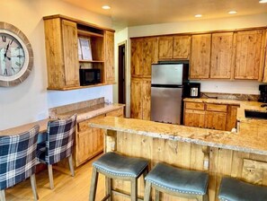 A kitchen with wooden cabinets, a stainless steel refrigerator, a microwave, and a clock on the wall. - Full Cabin Near Yellowstone & Teton National Parks (Ashton)