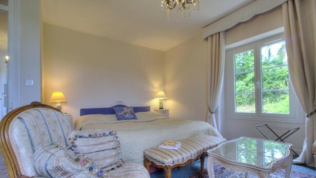 A bedroom with a bed, bedside lamps, a chair, a table, and a window with curtains. - The Cottage - ANDERT AND CONDON (ANDERT ET CONDON)