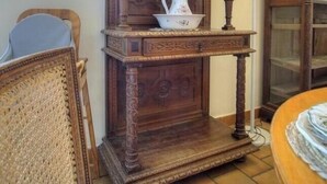 A room with a wooden cabinet, a wicker chair, and a table with a lamp. - The Cottage - ANDERT AND CONDON (ANDERT ET CONDON)