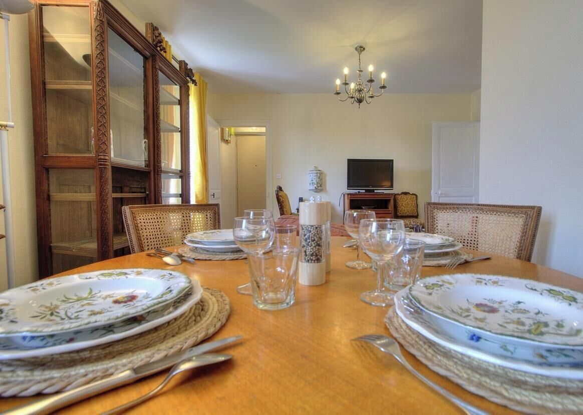 Une salle à manger avec une table en bois dressée pour quatre personnes, entourée de chaises en osier. Un téléviseur est fixé au mur, et un lustre est suspendu au plafond.