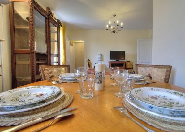 A dining room with a wooden table set for four, surrounded by wicker chairs. A television is mounted on the wall, and a chandelier hangs from the ceiling. - Valérie's Vacation Rental – ANDERT AND CONDON (ANDERT ET CONDON)