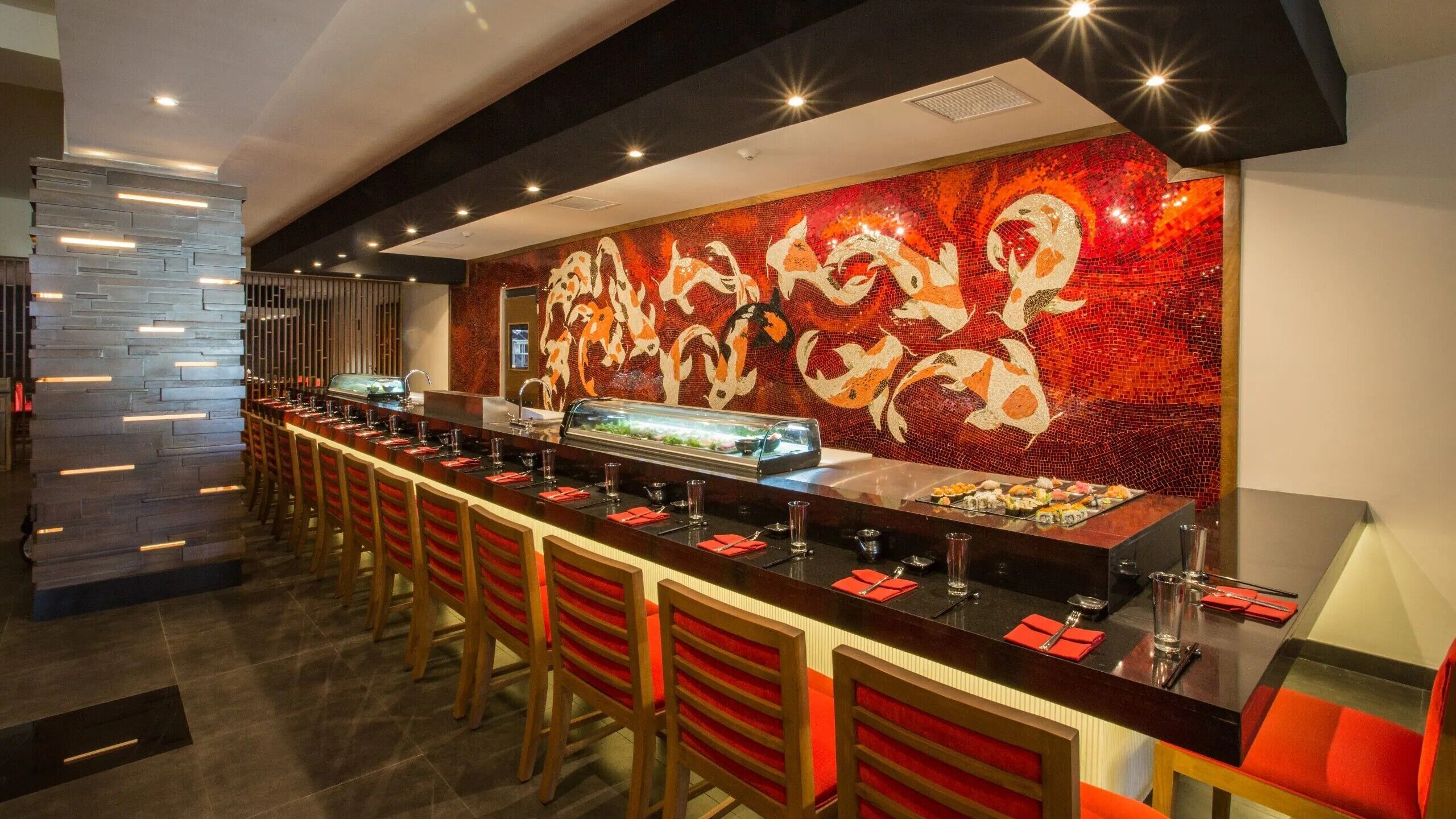A sushi bar with a long counter and red chairs, a decorative wall with koi fish mosaic, and a modern ceiling design.