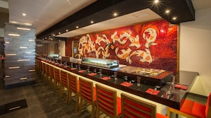 A sushi bar with a long counter and red chairs, a decorative wall with koi fish mosaic, and a modern ceiling design.