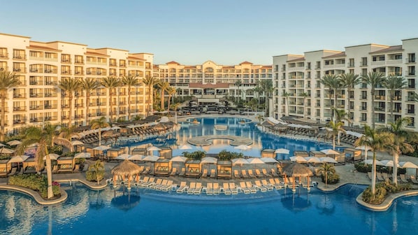 A resort with a central swimming pool surrounded by sun loungers and umbrellas, with multiple hotel buildings in the background.