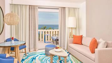 A hotel room with a balcony, a dining area with a round table and chairs, a sofa with cushions, and a view of the sea.