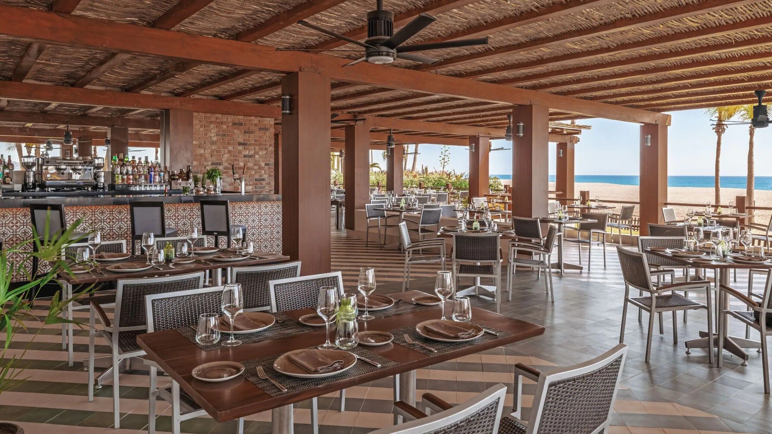 An outdoor restaurant with tables set for dining, a bar area with a variety of bottles, and a view of the beach.