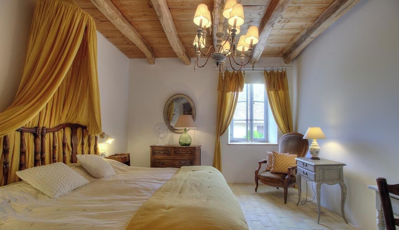 A bedroom with a bed, a wooden headboard, a chandelier, a dresser, a chair, and a window with curtains.