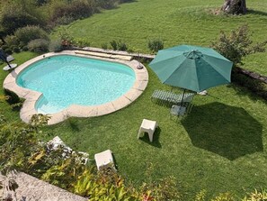 Een zwembad met een omliggende stenen rand, een groene parasol en twee witte stoelen.