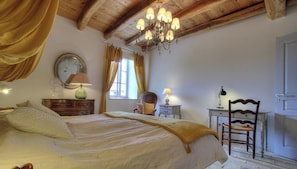 Een slaapkamer met een groot bed, een houten plafond, een kroonluchter, een dressoir, een stoel en een deur.