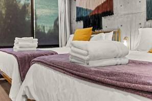 Ein Hotelzimmer mit zwei Betten, jedes mit weißer Bettdecke und lila Decke. Auf jedem Bett liegen Stapel weißer Handtücher.