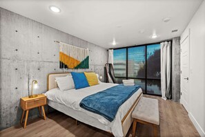 A modern bedroom with a large bed, a bedside table, a bench, and a view of the outdoors through a large window.