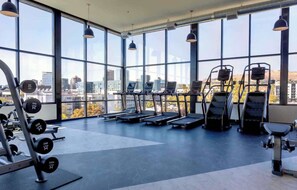 A gym with treadmills, weight machines, and dumbbells, large windows, and a city view.