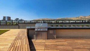 An outdoor barbecue area with a grill, wooden deck, and a view of a cityscape and hills. - Lux SLC - Pool - Gym - Rooftop (Salt Lake City)