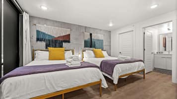 Quarto de hotel com duas camas, uma parede de concreto com tapeçaria colorida, piso de madeira e uma porta branca que dá para o banheiro.