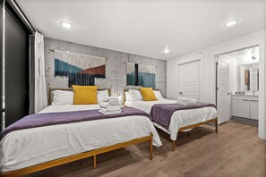 A hotel room with two beds, a concrete wall with a colorful tapestry, a wooden floor, and a white door leading to a bathroom.