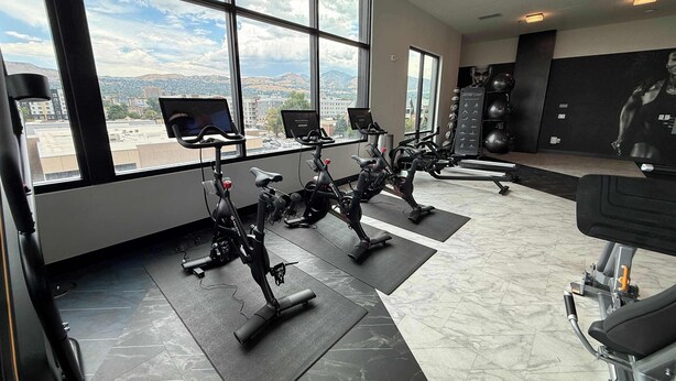 A gym with stationary bikes, exercise mats, and a large window with a city view. - Lux SLC - Pool - Gym - Rooftop (Salt Lake City)