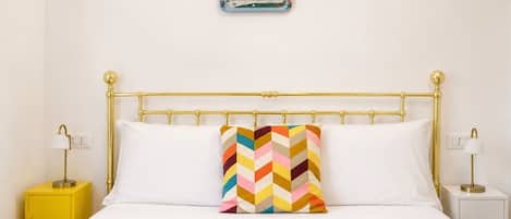 Una cama con estructura dorada, ropa de cama blanca y un cojín con diseño color chevron. Hay dos mesitas de noche, una con lámpara amarilla y otra con lámpara blanca. Encima de la cama, un pequeño marco azul cuelga en la pared.