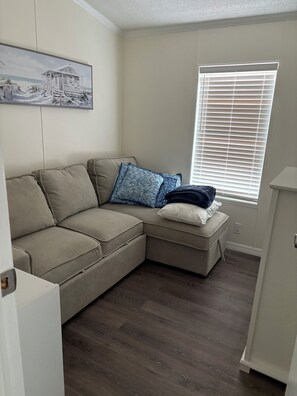 Ein Wohnzimmer mit einem beigen Ecksofa, einem kleinen Hocker, einem Fenster mit Jalousien und einem Bild an der Wand.