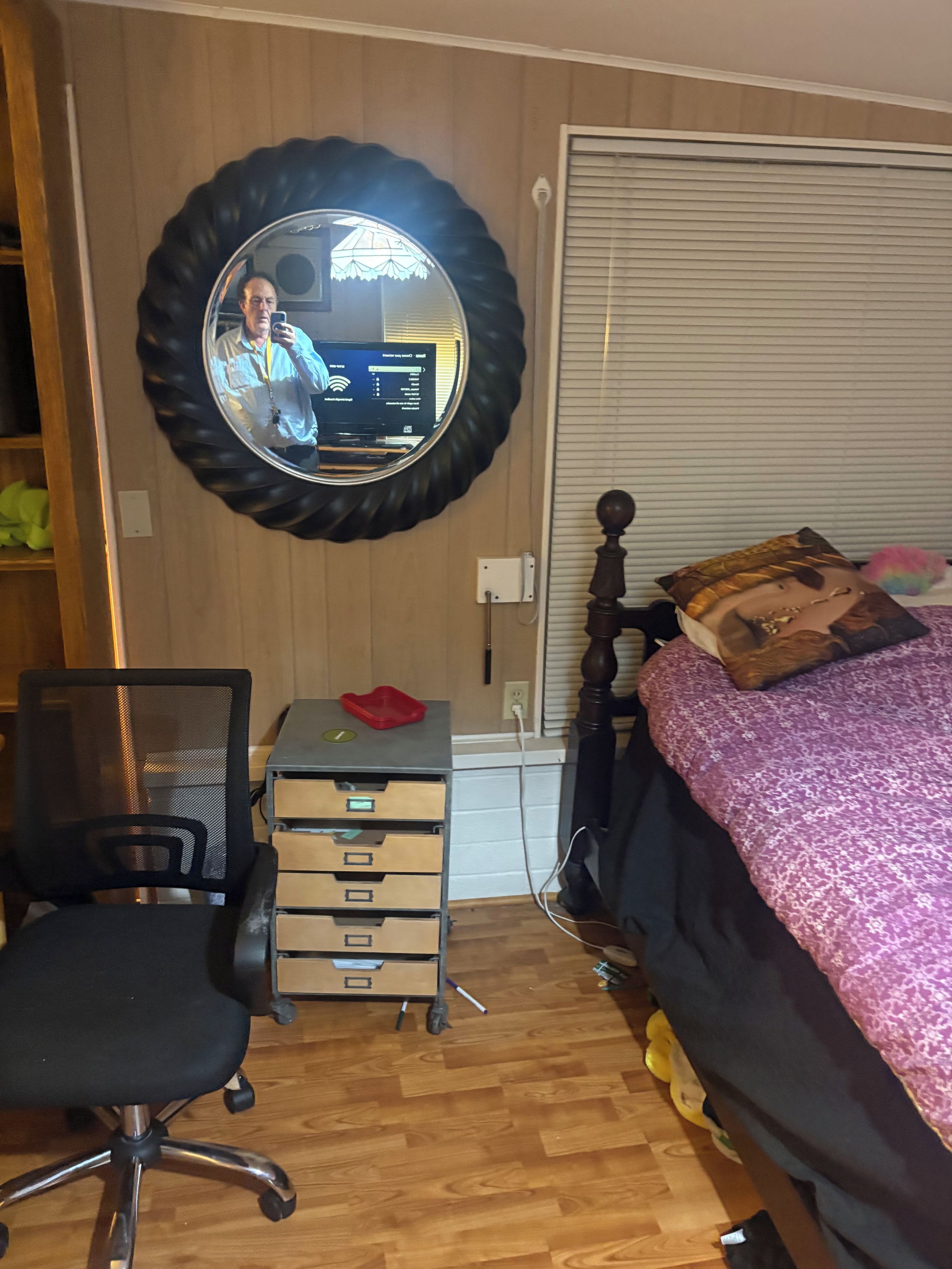 A bedroom with a bed, a desk with drawers, a chair, a mirror, and a window with blinds.
