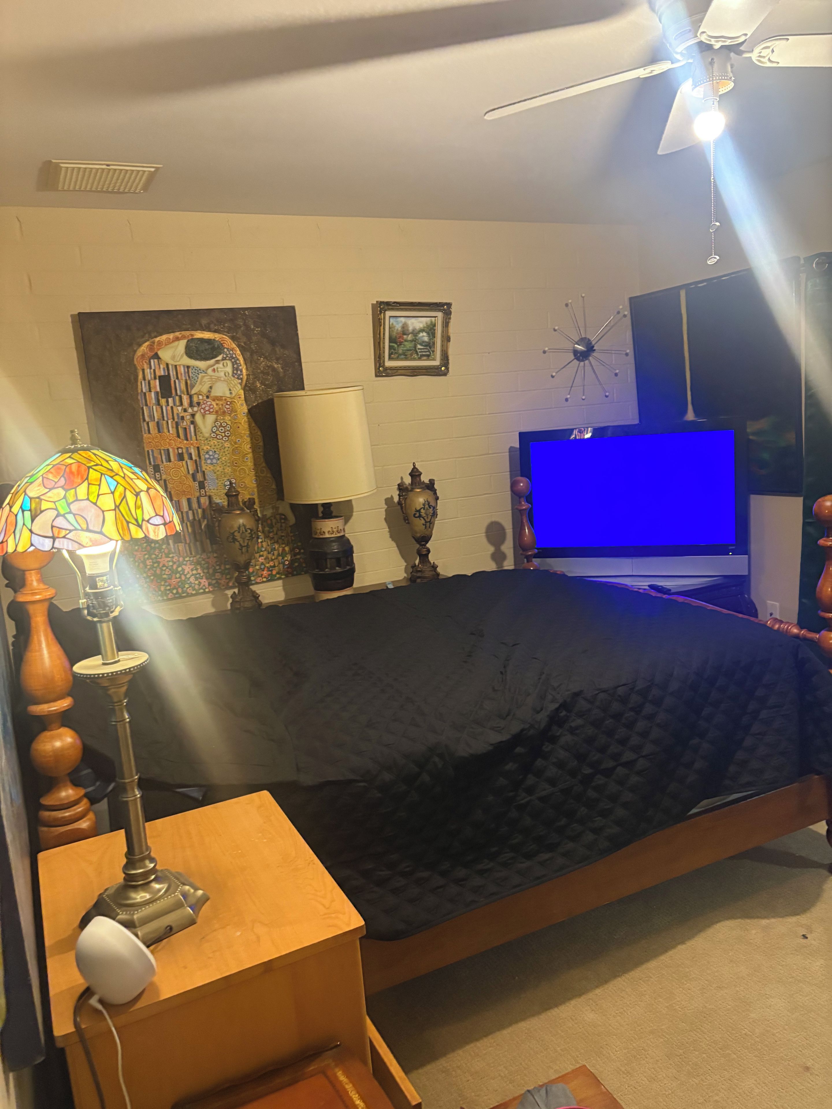 A bedroom with a bed, a desk with a lamp, a ceiling fan, a painting, and a TV.