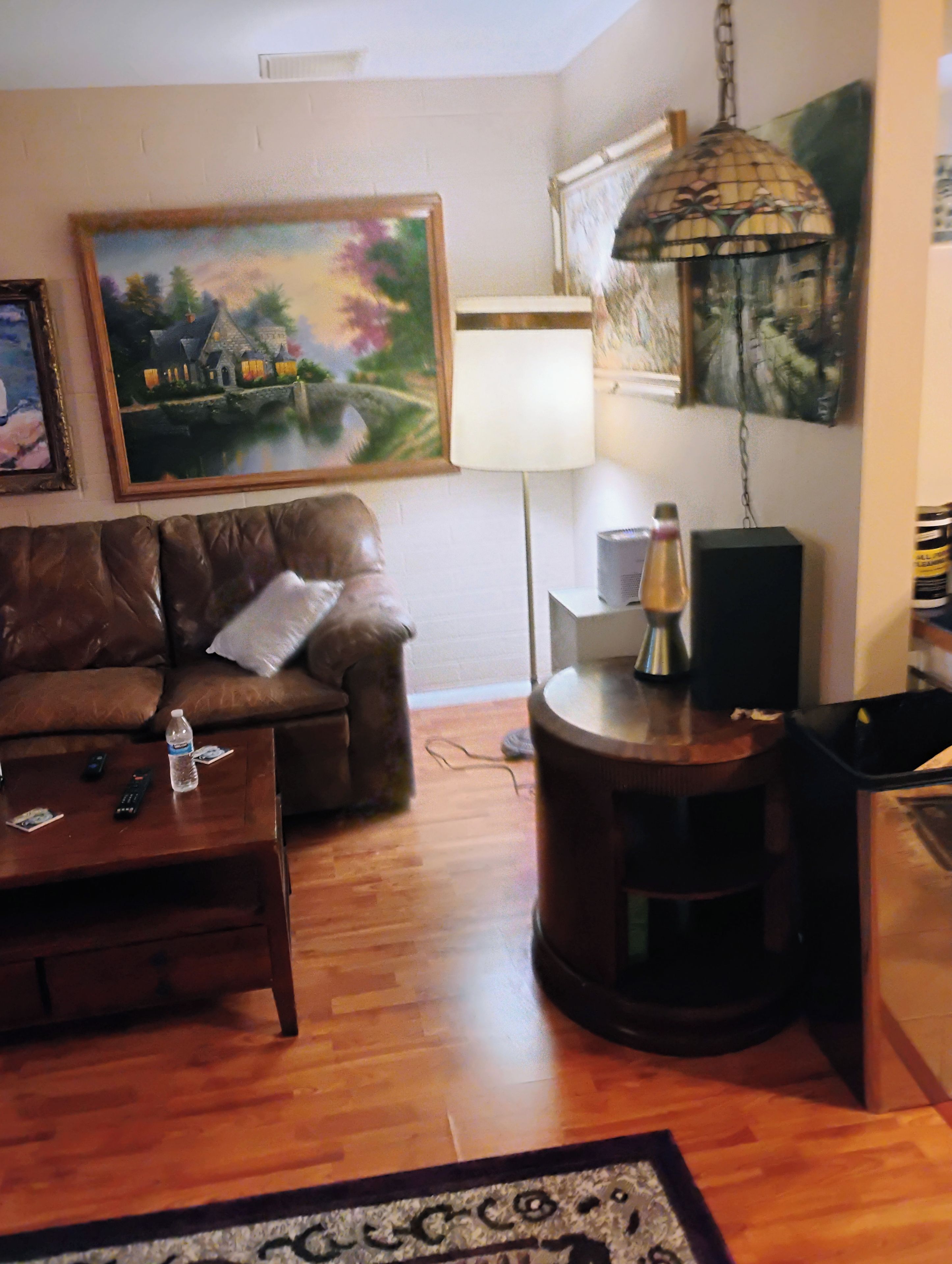 A living room with a brown leather sofa, a coffee table with a bottle and remote controls, a lamp, a speaker, and a patterned rug.