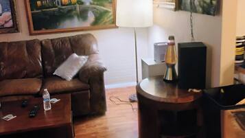 A living room with a brown leather sofa, a coffee table with a bottle and remote controls, a lamp, a speaker, and a patterned rug.