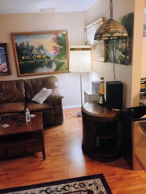 A living room with a brown leather sofa, a coffee table with a bottle and remote controls, a lamp, a speaker, and a patterned rug. - Spacious 4-bedroom house in serene Sun City with AC (Sun City)