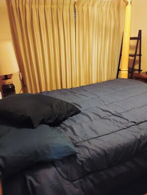 A bed with a dark blue bedspread and pillows, a lamp on a bedside table, and a curtain in the background. - Spacious 4-bedroom house in serene Sun City with AC (Sun City)
