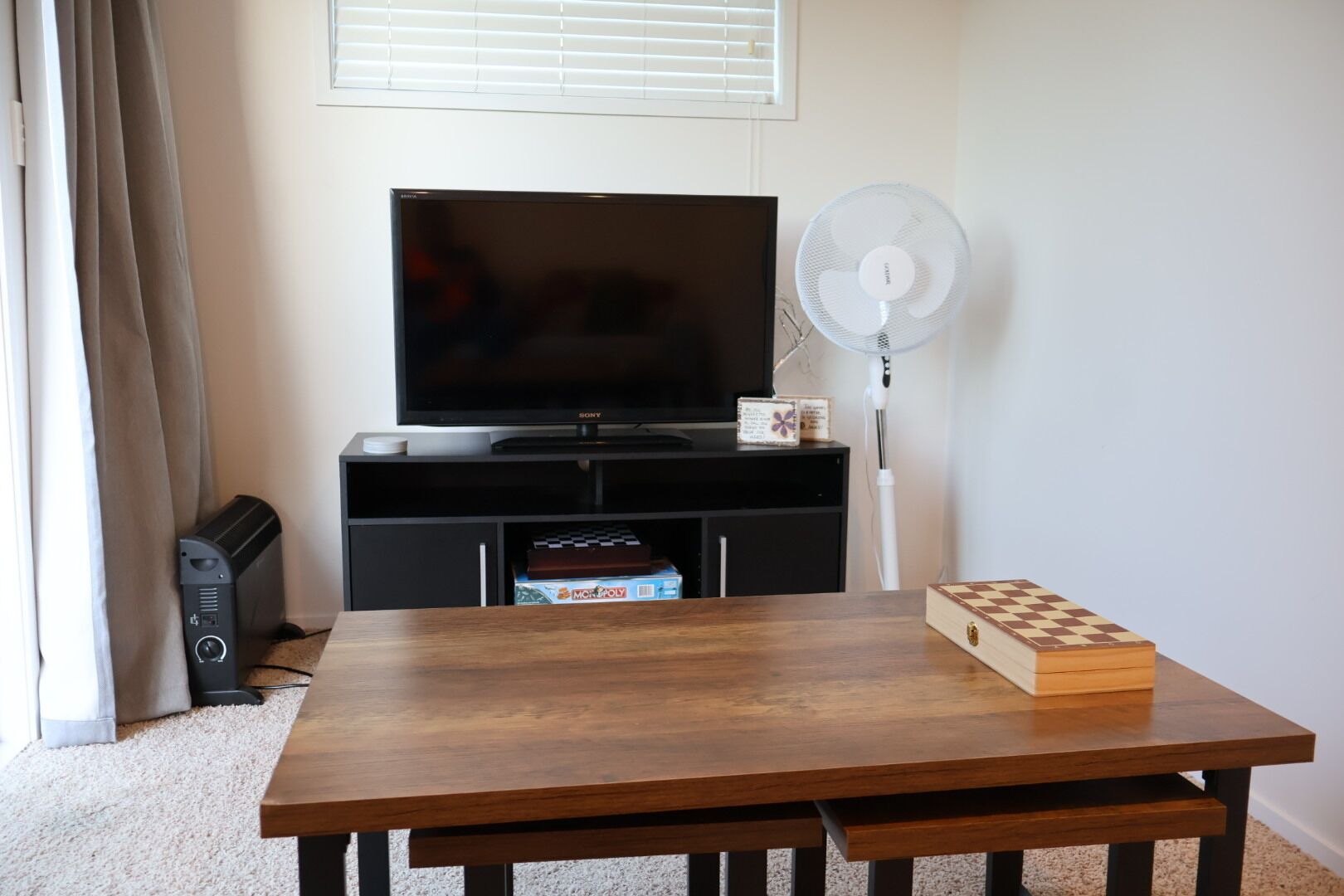 A living room with a flat-screen TV on a black stand, a white standing fan, a wooden table with a chessboard, and a carpeted floor.