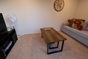 A living room with a wooden coffee table, a white wall clock, a white fan, a sofa with floral cushions, and a black entertainment unit.