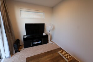 A living room with a black entertainment unit, a flat-screen TV, a standing fan, and a chessboard on the floor.