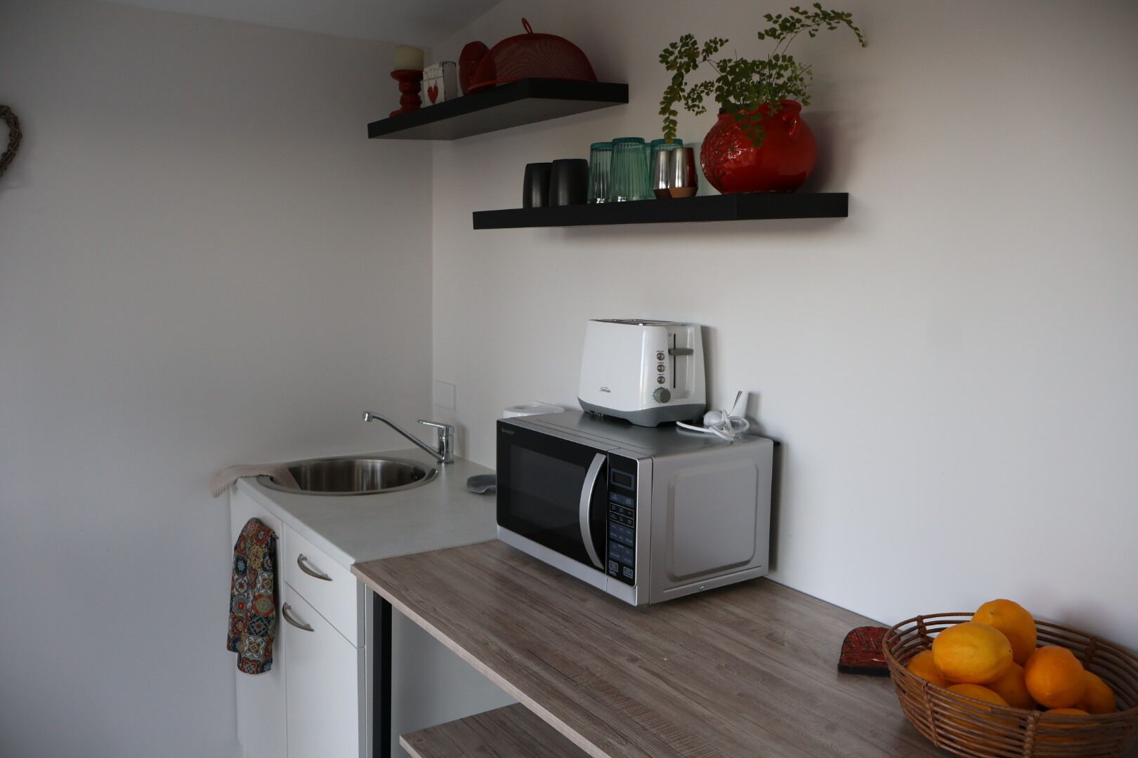 A compact kitchen with a microwave, toaster, and a sink. There are shelves above the counter holding various kitchen items and a basket of oranges on the counter.