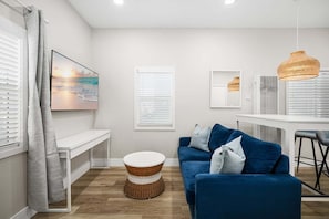 A modern living room with a blue sofa, a round wicker coffee table, and a large painting of a beach scene. - West Oceanfront Oasis 2-3-4-6-7 (Newport Beach)