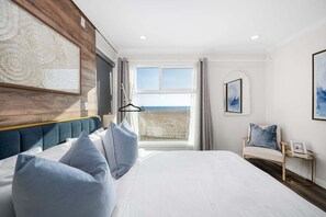 A bedroom with a large bed, a view of the beach, a chair, and a framed picture on the wall. - West Oceanfront Oasis 2-3-4-6-7 (Newport Beach)