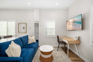 A modern living room with a blue sofa, a round wicker side table, a wooden desk with a chair, and a large flat-screen TV mounted on the wall. - West Oceanfront Oasis 1-5-8-9 (Newport Beach)