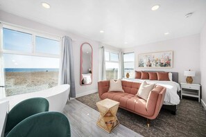 A hotel room with a large window, a bathtub, a sofa, a bed, and a painting. - West Oceanfront Oasis 1-5-8-9 (Newport Beach)