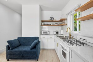 Eine kompakte Küche mit weißen Schränken, einem Gasherd, einer Mikrowelle und einer Kaffeemaschine. Im angrenzenden Wohnbereich befindet sich ein blaues Sofa mit Kissen.