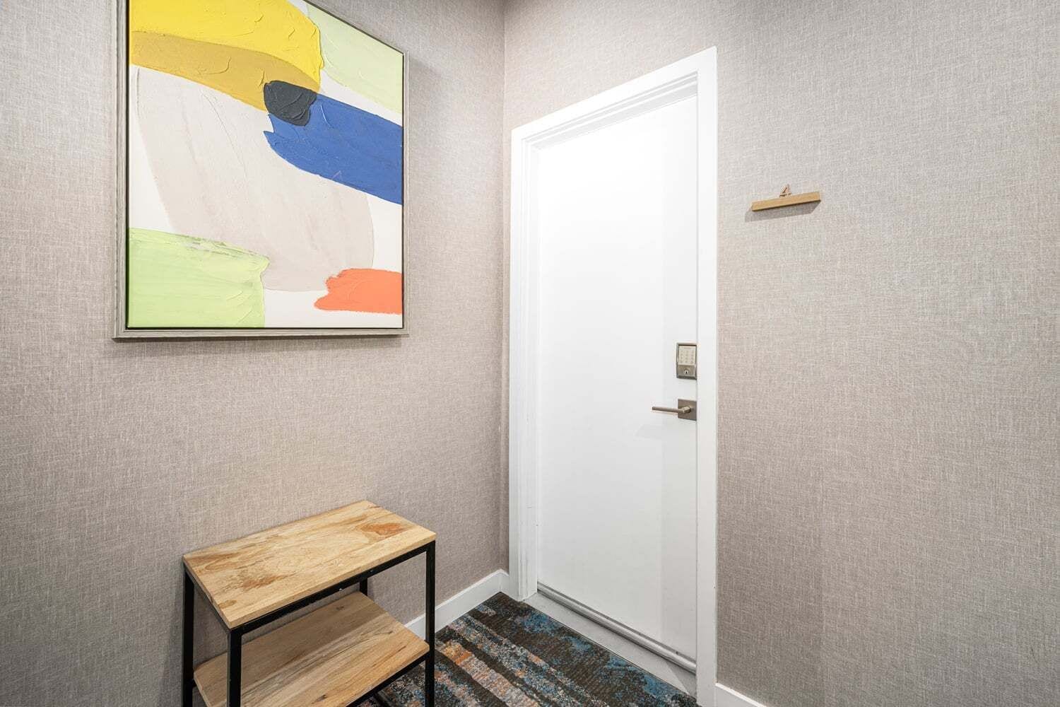 Un pasillo con una puerta blanca, una pequeña mesa de madera y un cuadro abstracto de colores en la pared.