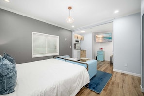A bedroom with a bed, a bedside table, a window with blinds, a kitchenette area, and a painting on the wall. - West Oceanfront Oasis 3-4-5-6-9 (Newport Beach)