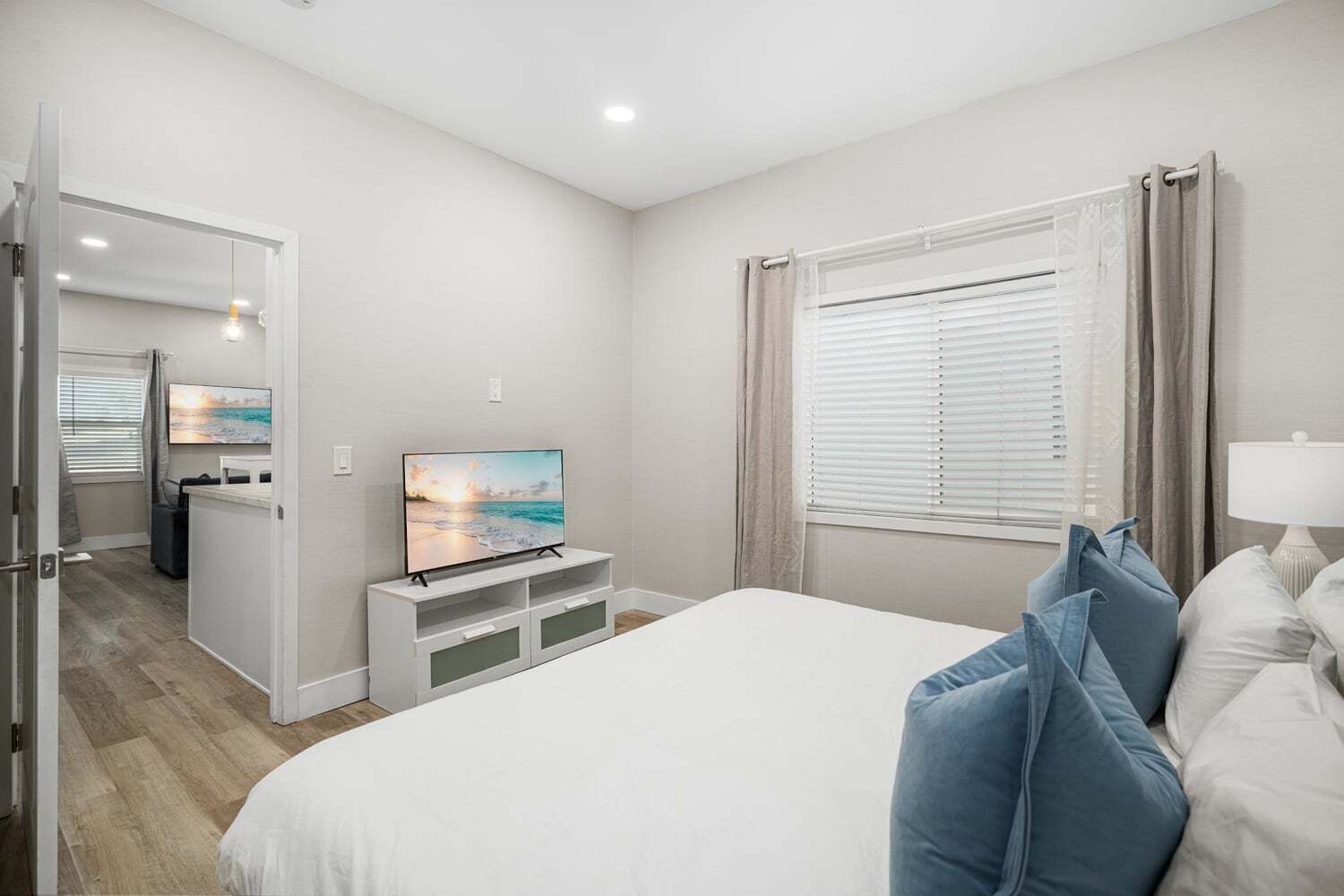 Habitación de hotel con cama, televisión sobre un mueble blanco, ventana con cortinas y puerta que da a otra habitación.