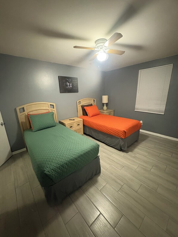A bedroom with two beds, a ceiling fan, a nightstand with a lamp, and a window with blinds.